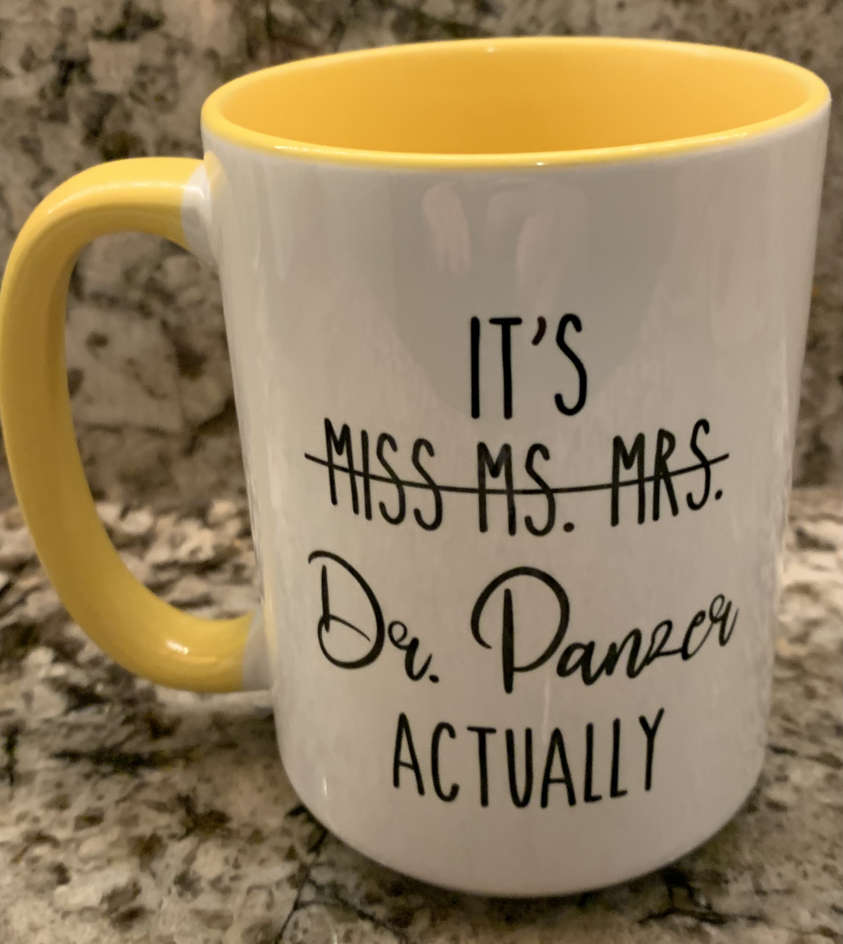 A large coffee mug with a sun-yellow handle and interior. The outside of the mug is white with black font. The words “Miss” “Ms.” and “Mrs.” are crossed out thus the text reads: “It’s Dr. Panzer actually”. Dr. Panzer is written in a fancier, cursive font to add emphasis.